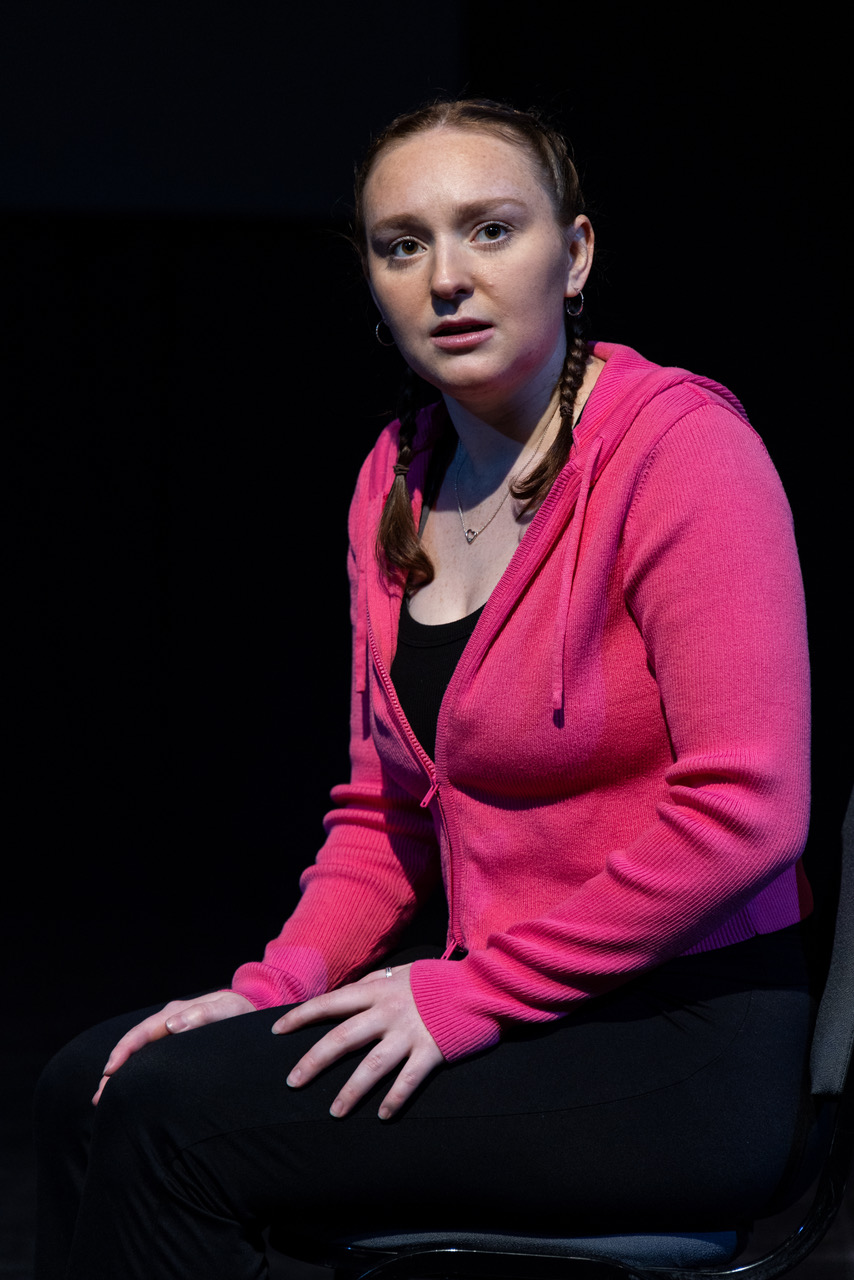 Ella, a white woman in her early 20s, siting in a black studio space. She is looking at something off to the right that we can't see, with an unclear expression on her face - is it concern?