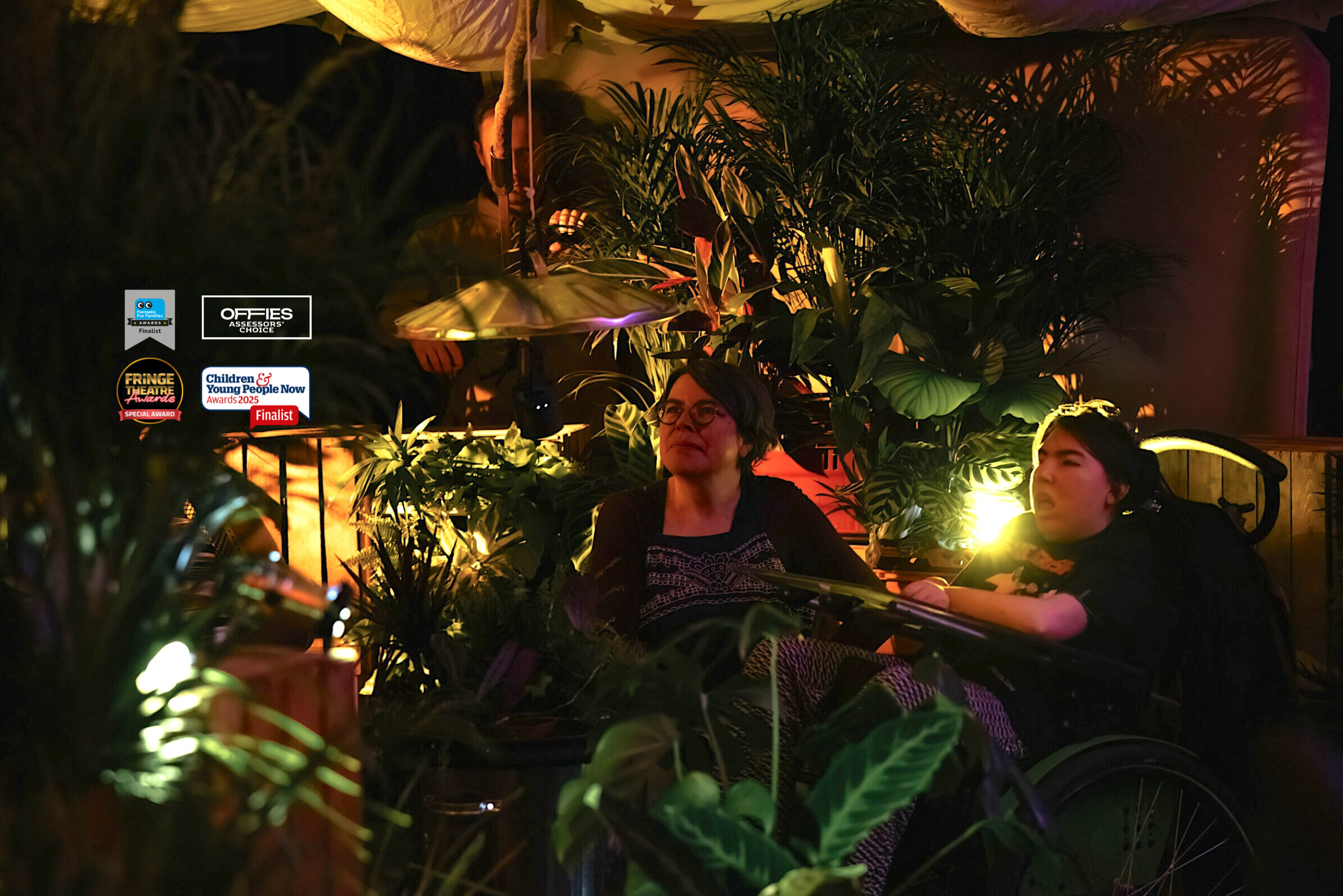A young woman with short brown hair sitting in her wheelchair. A middle aged woman with short grey hair and glasses sits next to her. They are inside the When the World Turns performance space, with a billowing tent overhead and surrounded with wooden crates and plants all around. There is atmospheric low lighting, illuminating their faces with a golden glow and casting shadows of the plant leaves. Hanging just above them is a metal bin lid. There are logos for awards and award shortlist places overlaid on the photo.