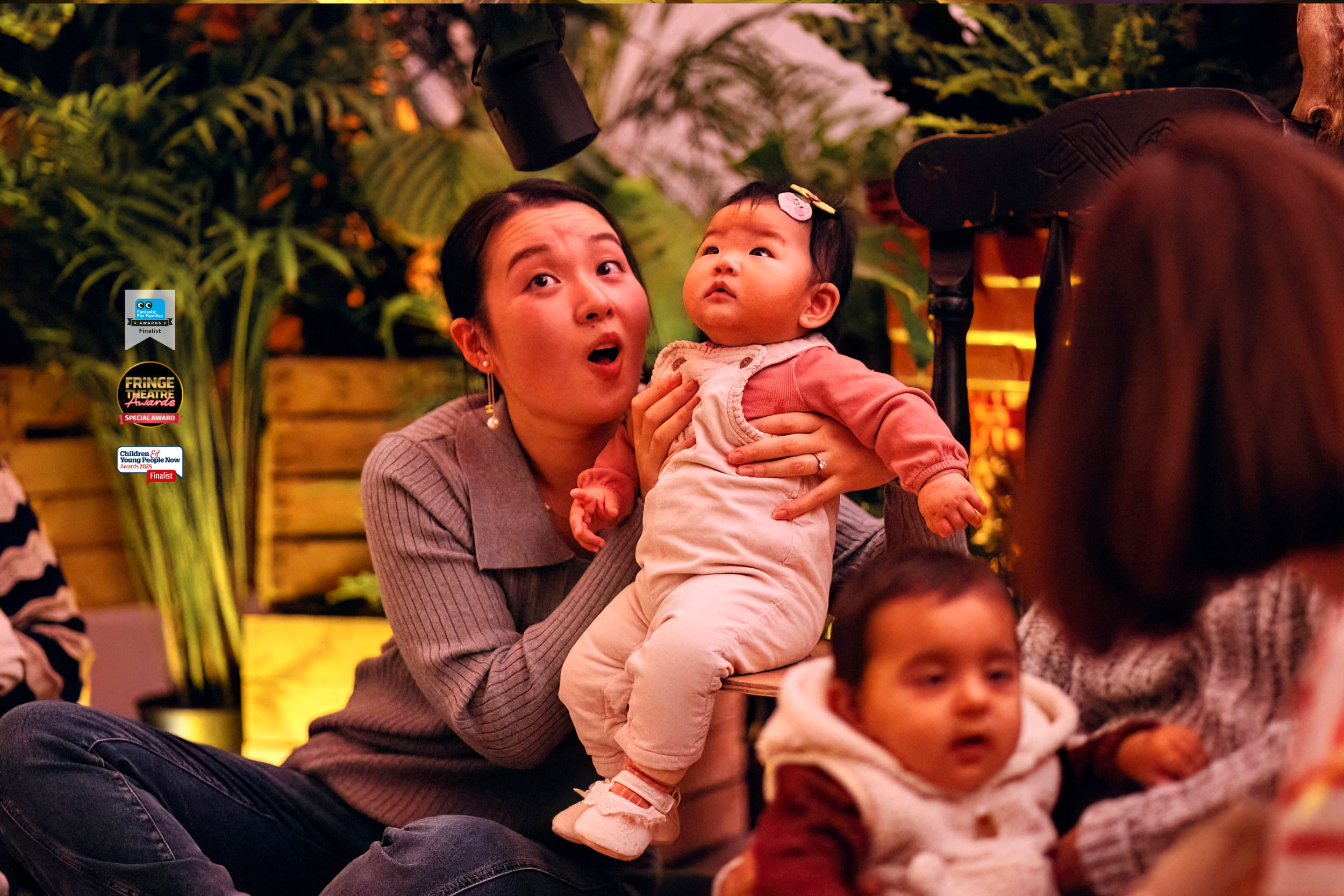 A mother sits on the floor, supporting a baby to sit on a chair next to her. They are both looking up at a speaker above them with wonder. Behind them are wooden crates filled with plants. There are logos for an award and award shortlist places overlaid on the photo.