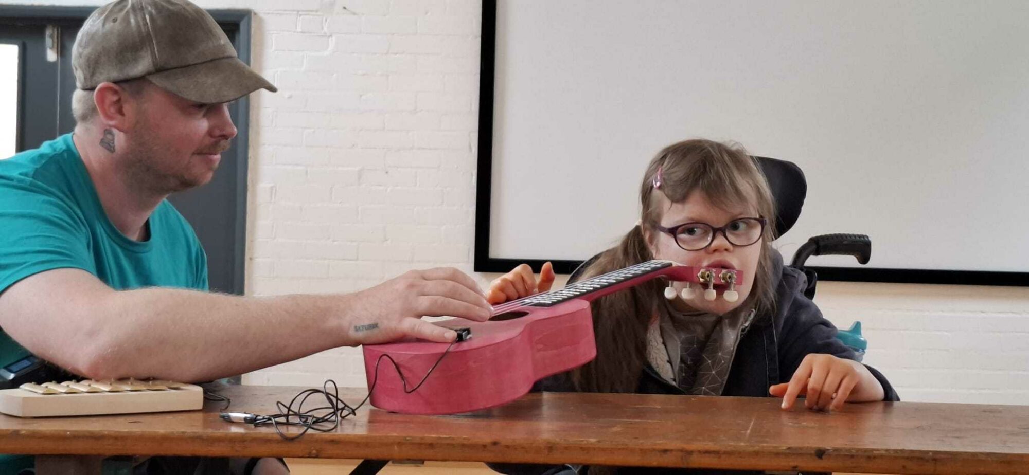 Youth Associate Lucy and InSideWays composer sit next to each other at a wooden table in a white studio space, during a creative jamming session for the show soundtrack. Lucy is a White female teenager with long, mid-brown hair and wears glasses. She is also a wheelchair user. She plays a pink guitar whilst holding the peg-head in her mouth. BK, an adult with a brown cap and teal t-shirt, looks on, one hand partly holding the guitar.