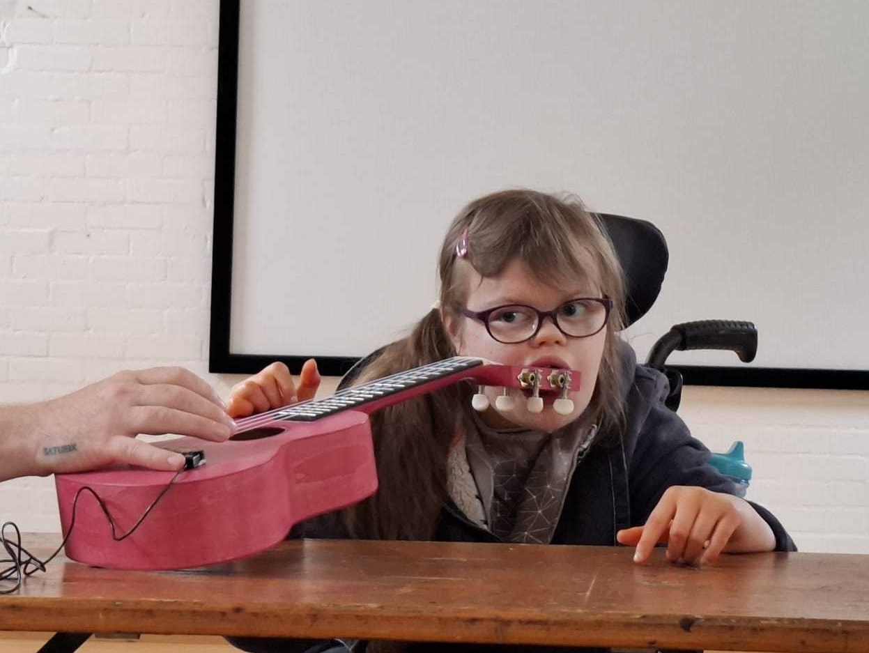 Youth Associate Lucy at a wooden table in a white studio space, during a creative jamming session for InSideWays. Lucy is a White female teenager with long, mid-brown hair and wears glasses. She is also a wheelchair user. She plays a pink guitar whilst holding the peghead in her mouth.