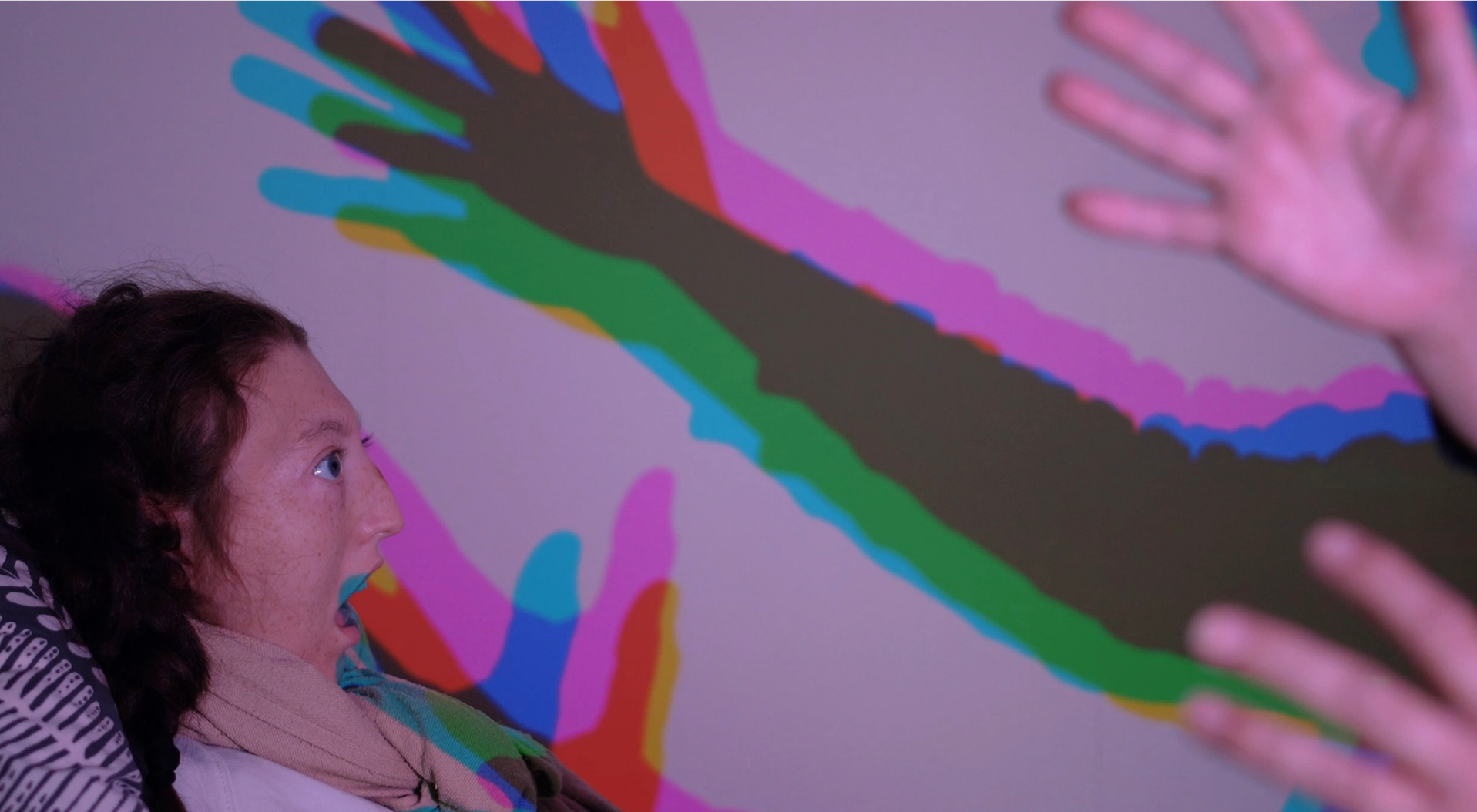 A young person looks up at hands held in front of them. They cast a colourful shadow on the walls.