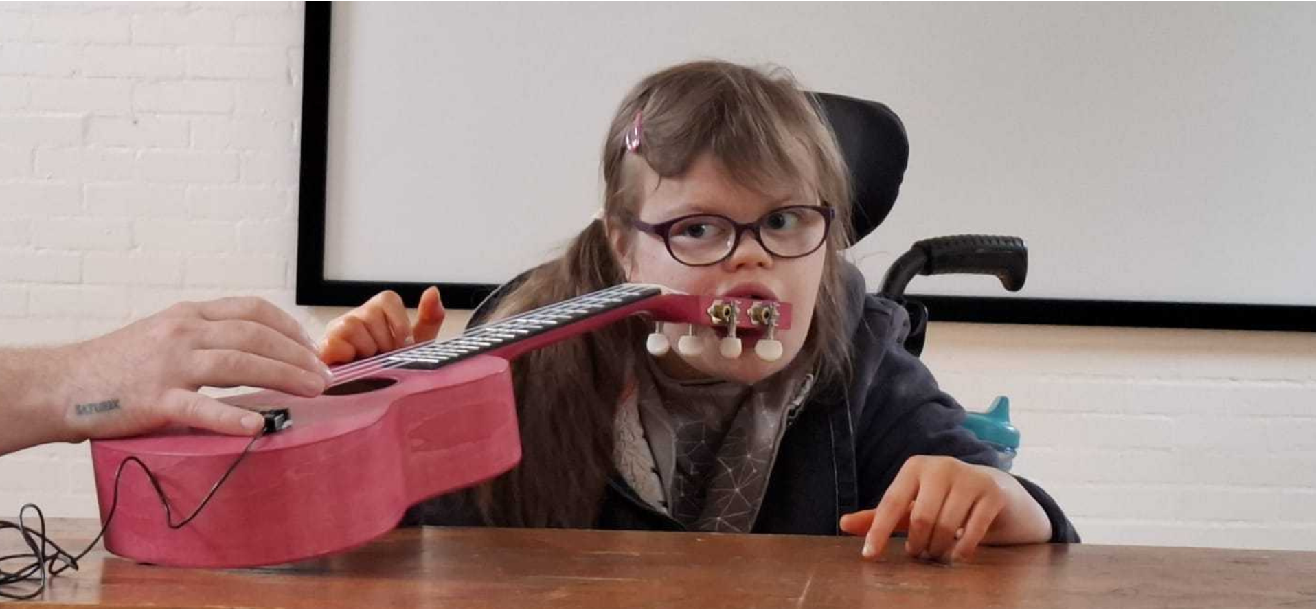 Youth Associate Lucy at a wooden table in a white studio space, during a creative jamming session for InSideWays. Lucy is a White female teenager with long, mid-brown hair and wears glasses. She is also a wheelchair user. She plays a pink guitar whilst holding the peghead in her mouth.