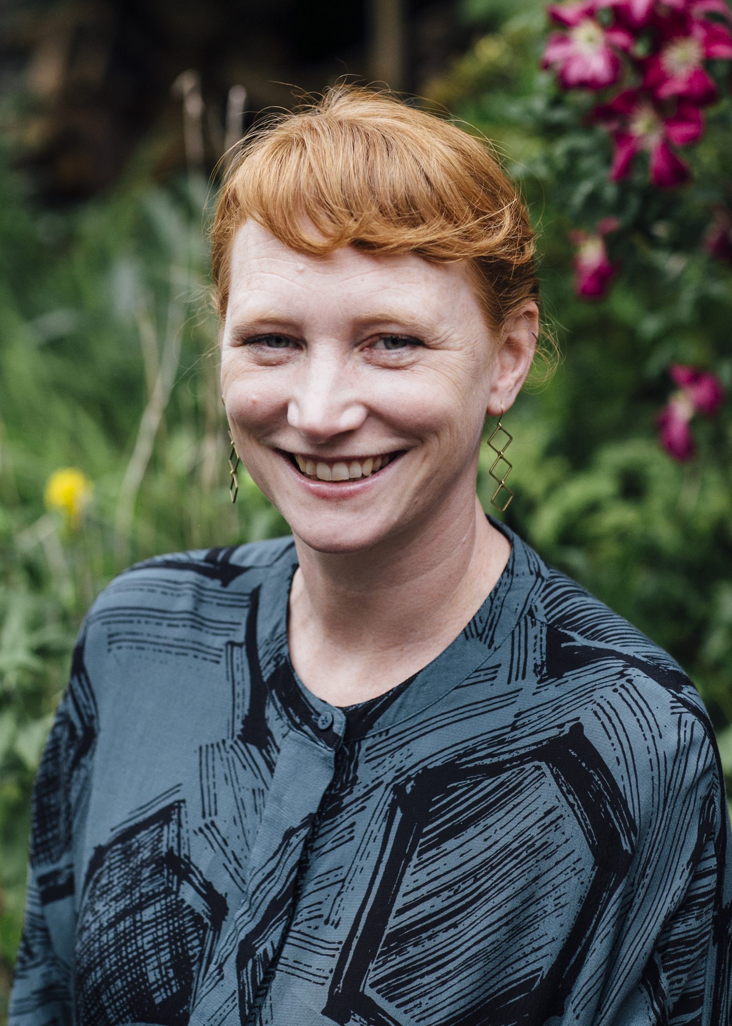 This photo shows Elspeth, a white woman in her 40’s sat in a garden wearing a green shirt. She has ginger hair and is smiling at the camera.
