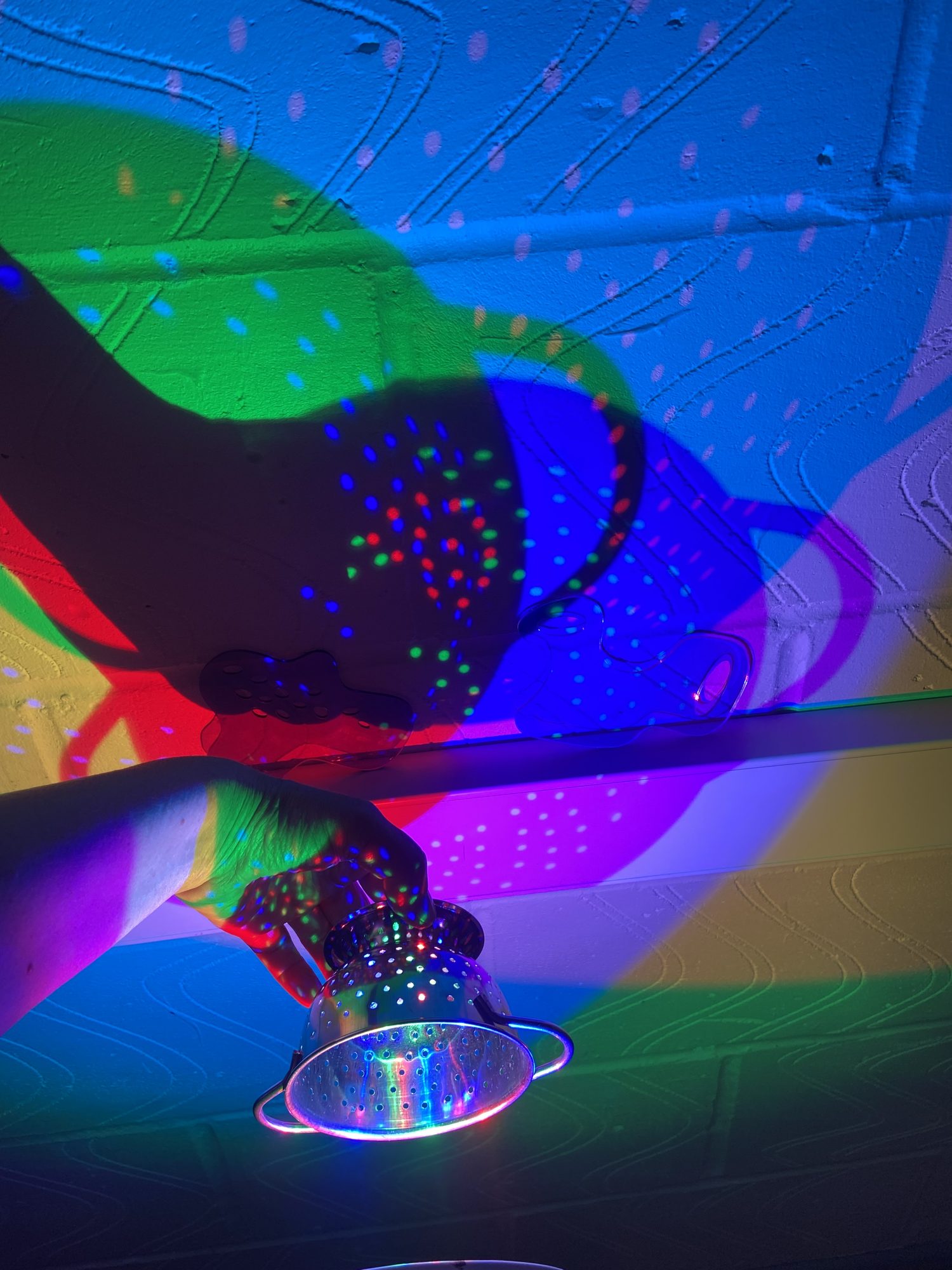 The background is blue and lots of overlapping bright colours and shapes. A hand is holding a small colander which is creating more interesting shadows.
