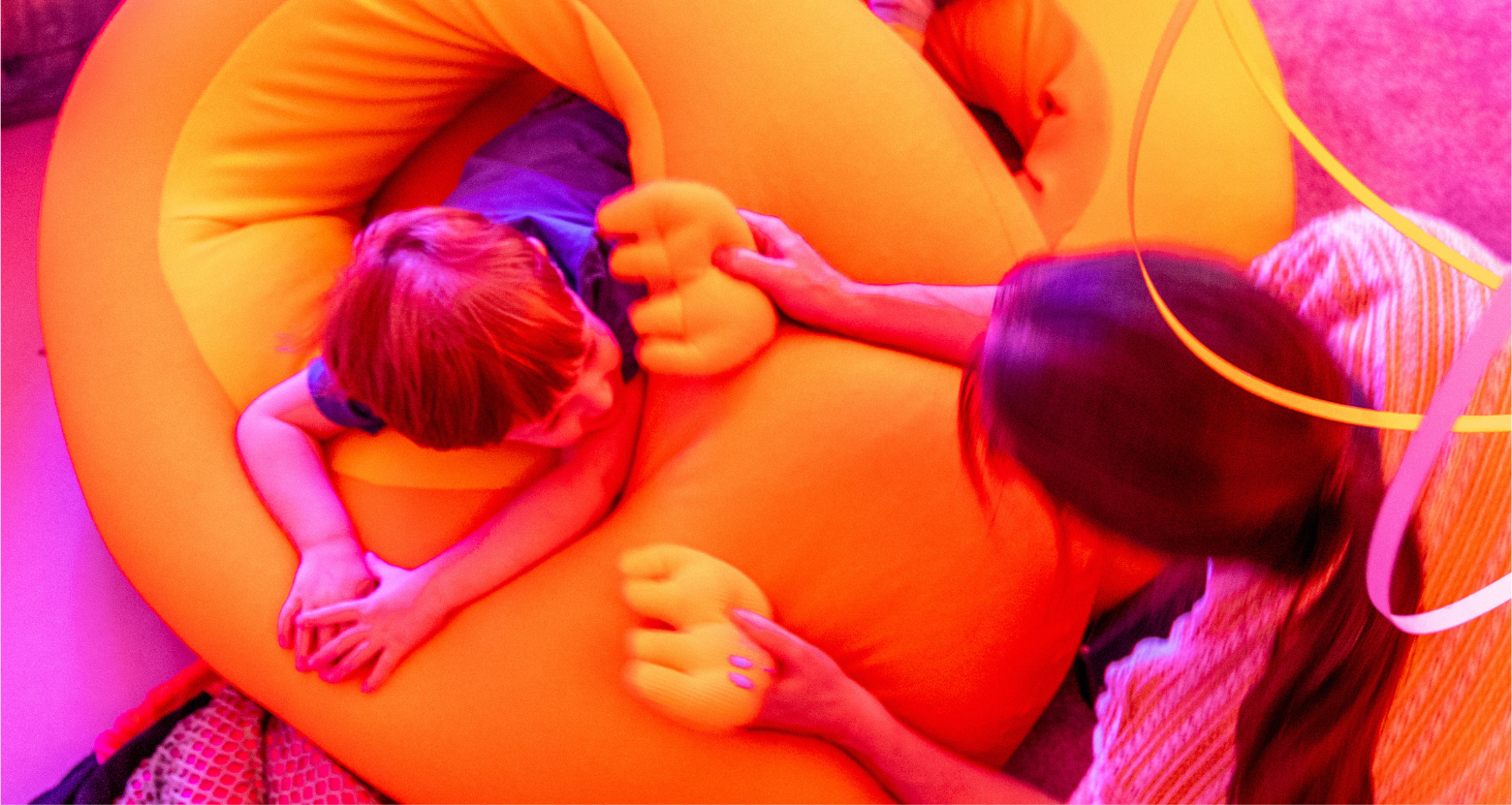 A child lies inside big, bright orange abstract stuffed shapes, that we call flumps. A parent holds out two little orange soft stuffed shapes that look a bit like elephants. Other colourful shapes and different textures surround them. Text reads: SideWays. A Sensory Playscape. 23 January - 6 June 2027. Royal Festival Hall. Southbank Centre.